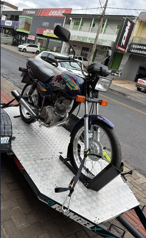 Frente da Titan azul em close: farol, guidão, tanque e roda dianteira, em rua urbana